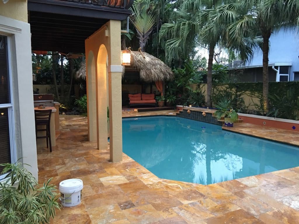 A pool framed by warm-toned paving, a covered patio, and tropical landscaping.